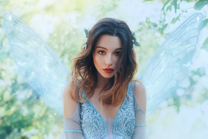 fantasy closeup portrait attractive woman in image young fairy. carnival costume blue dress transparent wings. face tender red makeup. wavy brunette hair flutter fly in wind. green nature backdrop