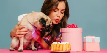 young woman petting dog and blowing candle on birthday cake while sitting at the pink desk on blue background