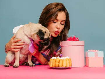 young woman petting dog and blowing candle on birthday cake while sitting at the pink desk on blue background